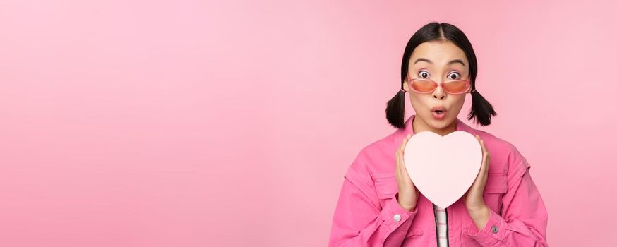 Beautiful asian girl smiling happy, showing heart gift box and looking excited at camera, standing over pink romantic background.