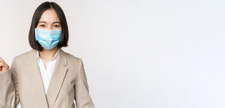 Coronavirus and work concept. Portrait of woman in medical face mask, pointing finger left, showing logo or banner, advertisement, white background.