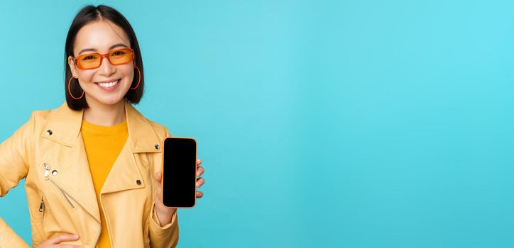 Image of young stylish asian female model in trendy sunglasses, showing mobile phone screen, application on smartphone interface, standing over blue background.