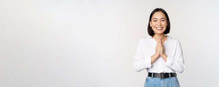 Portrait of happy asian girl laughing and smiling, showing thank you, namaste gesture, grateful for smth, standing over white background.