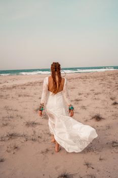 Model in boho style in a white long dress and silver jewelry on the beach. Her hair is braided, and there are many bracelets on her arms