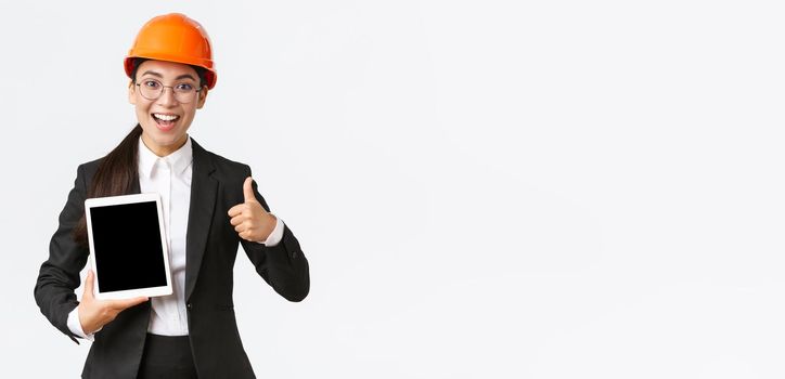 Impressed young female realtor, estate agent showing building during construction, wear safety helmet and business suit, holding digital tablet and make thumbs-up, recommend purchase.