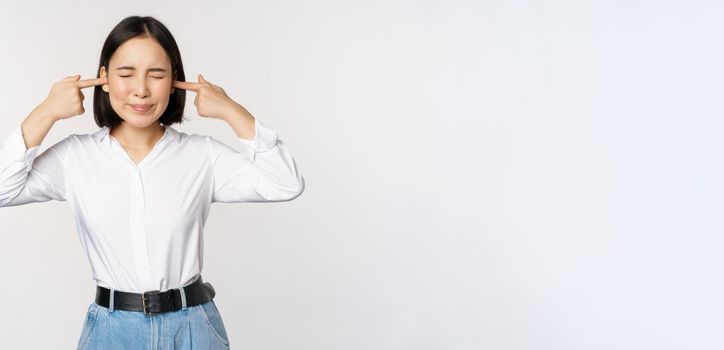 Annoyed asian woman feeling discomfort from loud annoying noise, close ears and eyes, standing over white background. Copy space