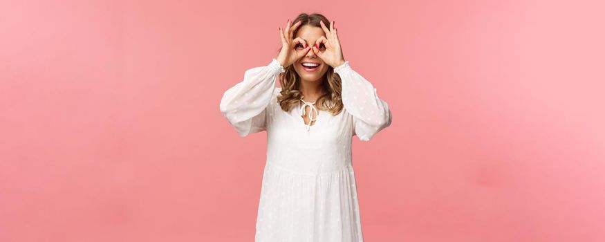Portrait of dreamy cute and funny young blond girl seeing something interesting, looking from okay signs as making glass-mask with fingers over eyes, smiling amused, pink background.