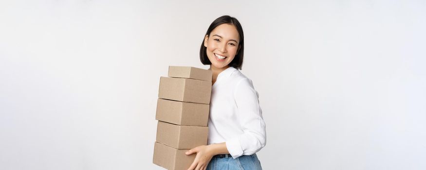 Portrait of young asian woman holding boxes, carry delivery goods. Korean female entrepreneur assemble order, standing voer white background.