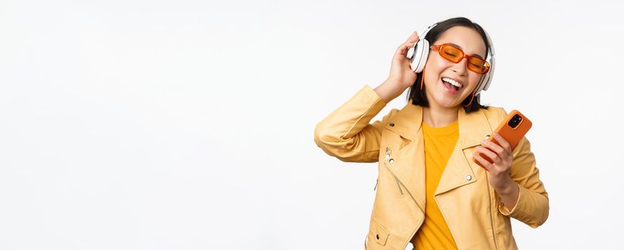 Technology concept. Stylish asian girl in headphones, holding smartphone, dancing and singing, listening music, standing over white background.