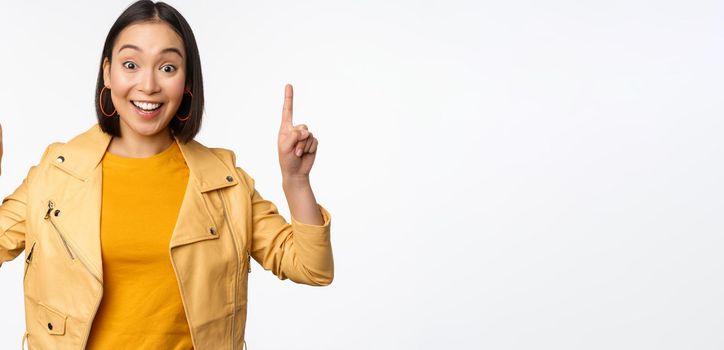 Enthusiastic asian girl pointing fingers up, showing advertisement on top, smiling happy, demonstrating promo offer or banner, standing over white background.
