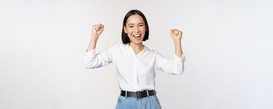 Enthusiastic asian woman rejoicing, say yes, looking happy and celebrating victory, champion dance, fist pump gesture, standing over white background.