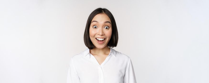 Image of korean woman looking surprised and happy at camera, standing over white background. Copy space