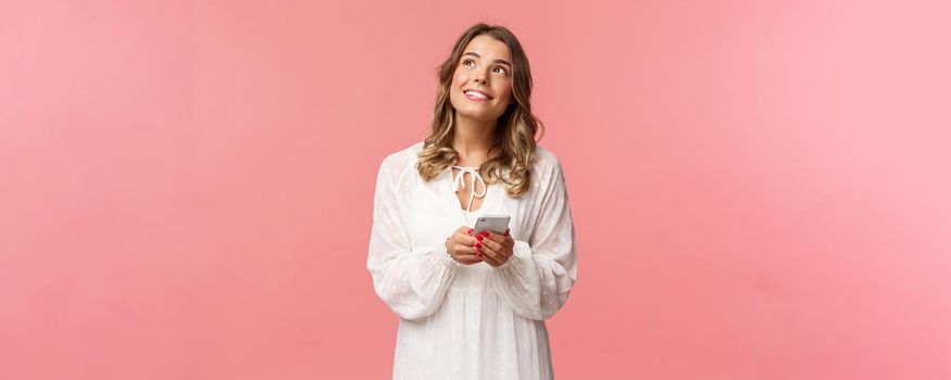 Portrait of dreamy young pretty blond girl fantasizing about something beautiful, looking up thoughtful and happy smiling, holding mobile phone, messaging, shopping online, pink background.