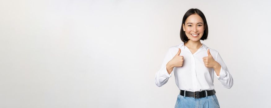 Image of beautiful adult asian woman showing thumbs up, wearing formal office, university clothing, recommending company, standing over white background.