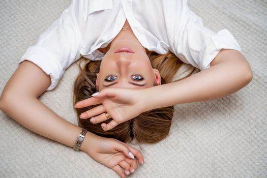 The woman lies on the bed on her back, top view. She looks straight ahead, wearing a white shirt.