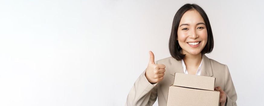 Smiling asian businesswoman, showing thumbs up and boxes with delivery goods, prepare order for client, standing over white background.