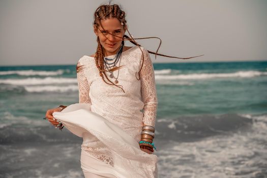 Model in boho style in a white long dress and silver jewelry on the beach. Her hair is braided, and there are many bracelets on her arms