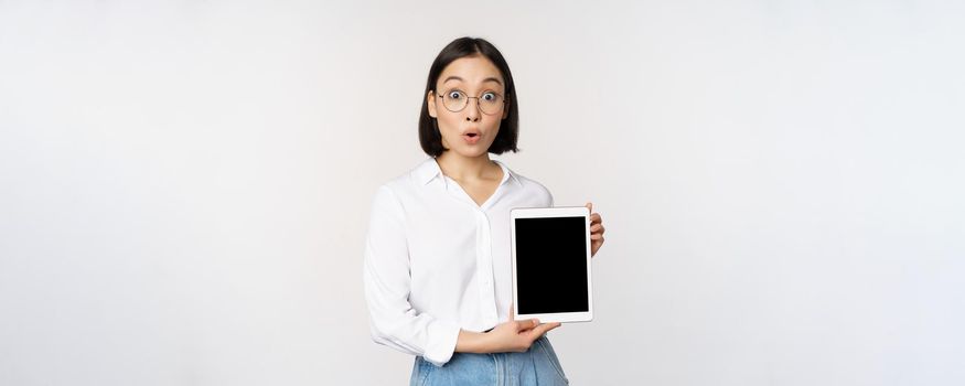 Enthusiastic asian woman, office worker in glasses showing digital tablet screen, demonstrating info on gadget display, standing over white background.