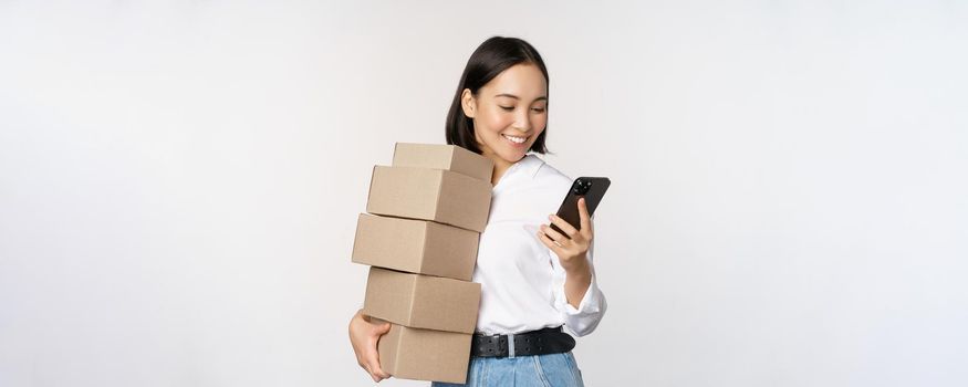 Image of young asian woman holding boxes, customer orders and looking at mobile phone, standing over white background.