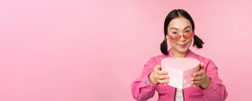 Cute asian girl giving you gift in heart shaped box, kissing and smiling, concept of holiday and celebration, standing over pink background.