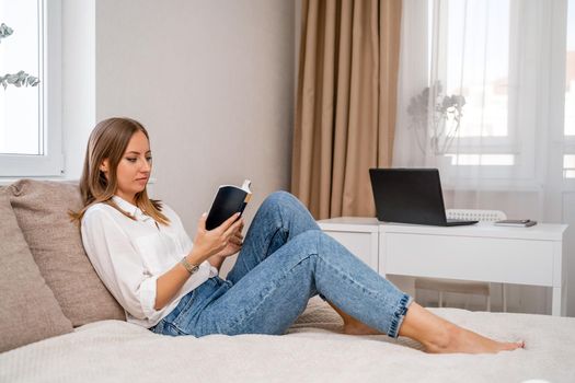 White cozy bed and a beautiful girl in a white shirt and jeans reading a book, the concept of home and comfort. Against the background of the living room, computer and window