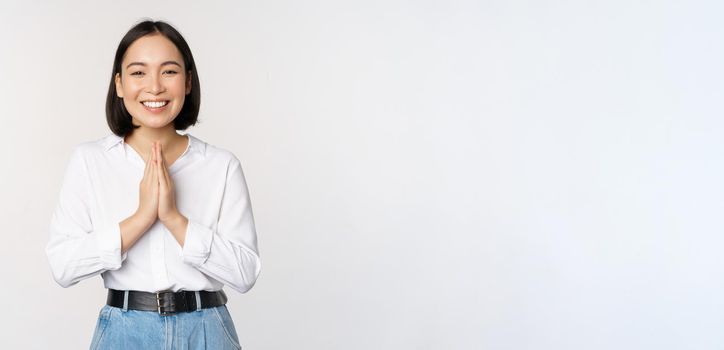 Portrait of happy asian girl laughing and smiling, showing thank you, namaste gesture, grateful for smth, standing over white background.