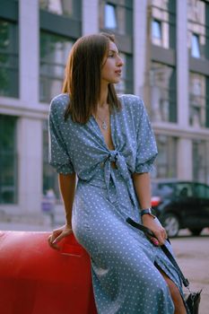 Cute blonde maiden in a long blue dress with polka-dots, watch, with a pendant around her neck and a small black handbag on her shoulder is looking away, posing sitting on a red guardrail while walking alone in the city. The concept of fashion and style. Close-up shot.