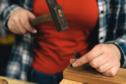 Detail of the hands of a young woman carpenter with red hair, concentrated and precise, working on the design of wood in a small carpentry workshop, dressed in a blue checked shirt and a red t-shirt. Detail of a young woman carpenter's hands holding a nail in a wooden board. Warm light indoors, background with wooden slats. Horizontal.