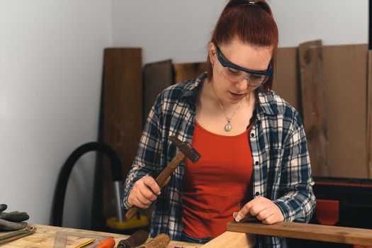 Detail of the hands of a young red-haired carpenter woman, concentrated and precise, working on the design of wood in a small carpentry workshop, dressed in a blue checked shirt and a red t-shirt. Carpenter woman holding a nail in a wooden board, in her small carpentry business. Warm light indoors, background with wooden slats. Horizontal.