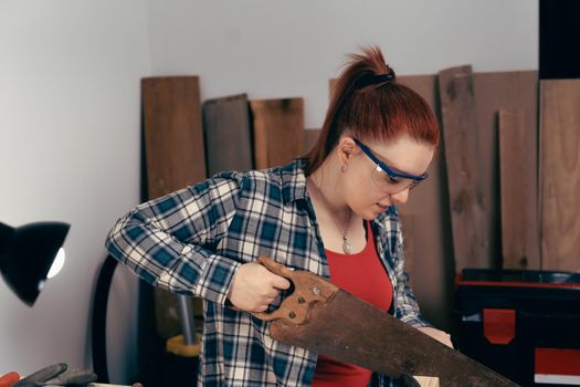 young red-haired female carpenter, concentrated and precise, working on the design of wood in a small carpentry workshop, dressed in blue checked shirt and red t-shirt. Young businesswoman sawing a wooden board in her small workshop and designing new furniture for the house. Warm light indoors, background with wooden slats. Horizontal.