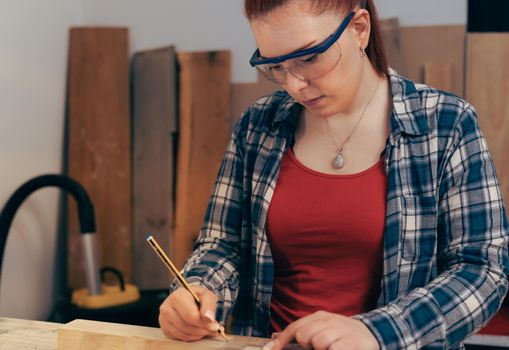 Young red haired carpenter woman, concentrated and precise, working on wood design in a small carpentry workshop, dressed in blue checked shirt and red t-shirt. Young businesswoman measuring a wooden board in her small workshop and designing new furniture for the house. Warm light indoors, background with wooden slats. Horizontal.