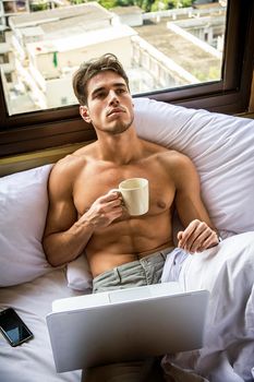 Naked young man with muscular body on bed with mug or cup in hand with coffee or tea, looking at laptop computer screen