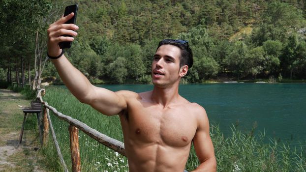 Young shirtless muscular handsome man doing videochat with smartphone at lake in forest