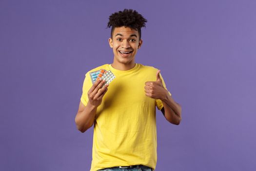 Health, influenza, covid-19 concept. Portrait of young healthy man got better, show thumbs-up holding drugs, taking pills to feel better after catching cold, being sick, purple background.