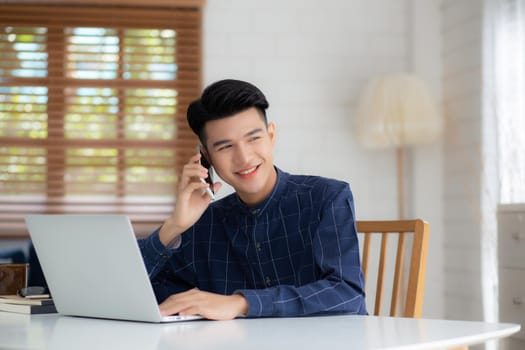 Young asian man talking phone and work from home with laptop computer, freelance using notebook to internet online and speak on smartphone, domestic life, business and communication concept.