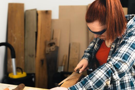 Close-up cropped shot of a young woman carpenter with red hair, concentrated and precise, working on wood design in a small carpentry workshop, dressed in blue checkered shirt and red t-shirt. young woman amateur carpenter, repairing an antique piece of furniture in her antique house. Warm light indoors, background with wooden slats. Horizontal.