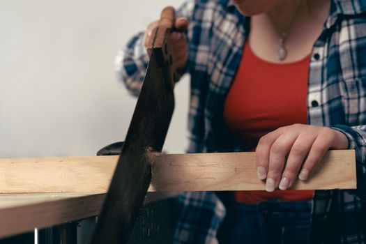 Close up cropped shot of a young red haired female carpenter, concentrated and precise, working on wood design in a small carpentry workshop, dressed in blue checked shirt and red t-shirt. Young businesswoman sawing a wooden board in her small workshop and designing new furniture for the house. Warm light indoors, background with wooden slats. Horizontal.