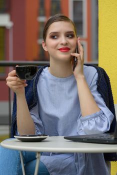 Charming happy woman student communicates by phone and use laptop computer to prepare for the course work. Concept of working outdoors