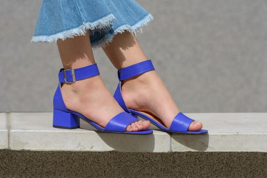 Women's legs in blue denim jeans and sandals in the city street. Trendy elegant casual outfit. Details of everyday summer look. Street fashion.