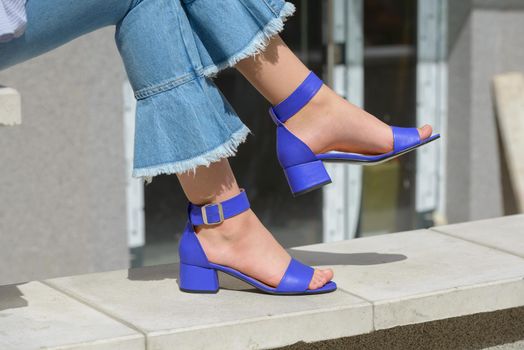 Women's legs in blue denim jeans and sandals in the city street. Trendy elegant casual outfit. Details of everyday summer look. Street fashion.