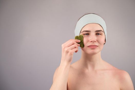 Portrait of a young woman massages her face with a gouache scraper on a white background