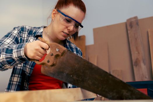 Close-up of a young red-haired female carpenter, concentrated and precise, working on the design of wood in a small carpentry workshop, dressed in blue checked shirt and red t-shirt. Young businesswoman sawing a wooden board in her small workshop and designing new furniture for the house. Warm light indoors, background with wooden slats. Horizontal.