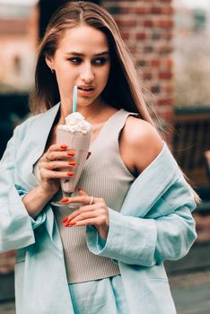 A woman's hand holds a refreshing cocktail in a glass with a tube. tanned woman wearing a beige T-shirt and a blue cotton jacket. Orange manicure