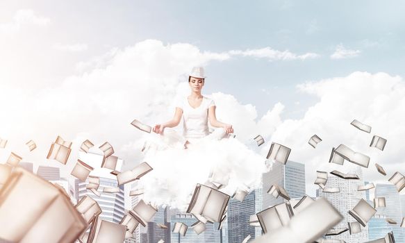 Woman in white clothing keeping eyes closed and looking concentrated while meditating on cloud among flying books with cityscape view on background.
