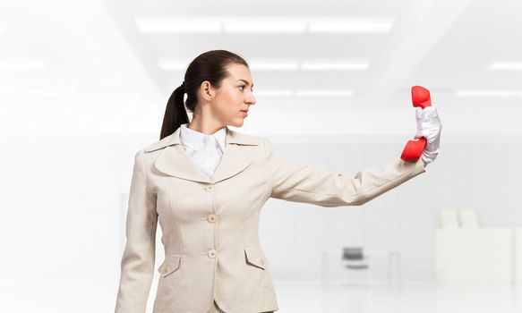 Woman keep at distance vintage red phone. Elegant operator in business suit posing with telephone in light office interior Hotline telemarketing and communication. Business assistance and support