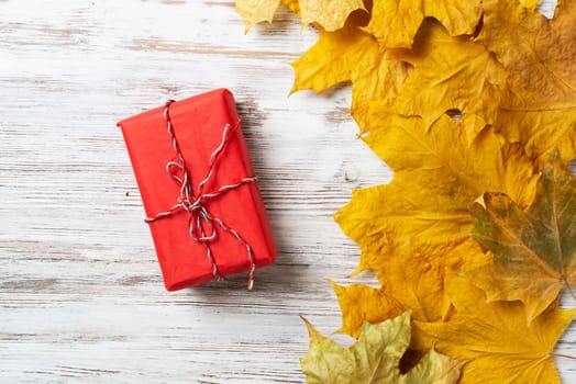 Bright autumn composition with gift box and yellow maple leaves. Holiday present in red wrapping paper lies on vintage wooden desk. Happy thanksgiving congratulation. Autumn sale advertising.