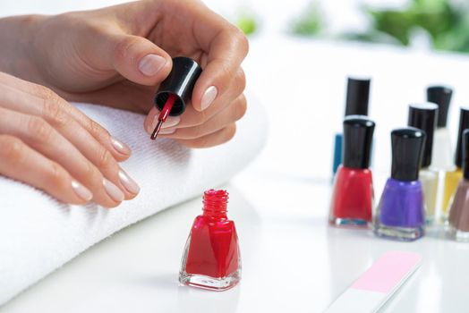 Woman giving herself elegant manicure at home. Closeup of beautiful female hand applying red nail polish. Colorful nail polish bottles on table. Nail care and beautician procedure. Stylish nail art