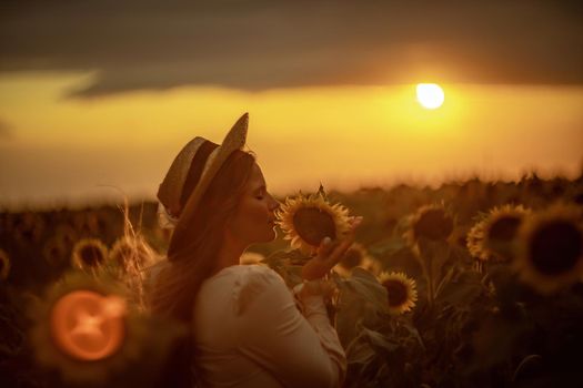 Beautiful middle aged woman looks good in a hat enjoying nature in a field of sunflowers at sunset. Summer. Attractive brunette with long healthy hair