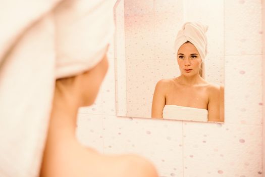 Portrait of young girl with towel on head in white bathroom looks and touches her face in the mirror and enjoys youth and hydration. Natural beauty, home care for problem skin.