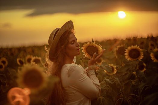 Beautiful middle aged woman looks good in a hat enjoying nature in a field of sunflowers at sunset. Summer. Attractive brunette with long healthy hair