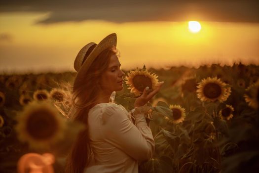 Beautiful middle aged woman looks good in a hat enjoying nature in a field of sunflowers at sunset. Summer. Attractive brunette with long healthy hair