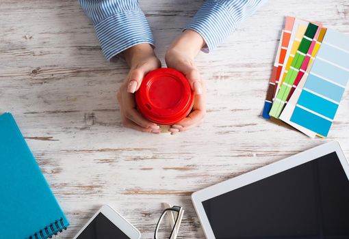 Office manager holding cardboard cup of coffee at wooden desk. Office workspace with tablet computer and accessories. Coffee break at office workplace. Young woman drinking coffee from disposable cup