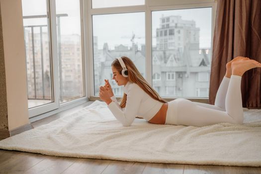 Side view portrait of relaxed woman listening to music with headphones lying on carpet at home. She is dressed in a white tracksuit